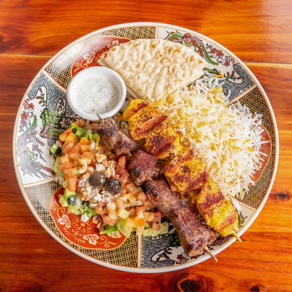 Two skewers of grilled meat, rice, salad, pita bread, and a dipping sauce on a patterned plate.
