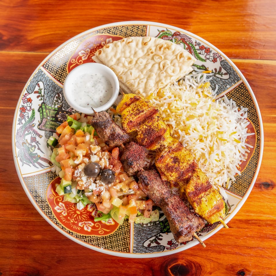 Two skewers of grilled meat, rice, salad, pita bread, and a dipping sauce on a patterned plate.