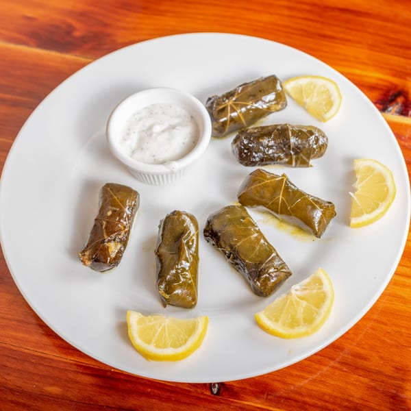 Six dolmades, a small bowl of white sauce, and lemon wedges on a white plate.
