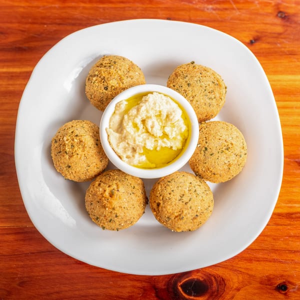 Six golden-brown falafel balls surround a white bowl of hummus drizzled with olive oil on a white plate.