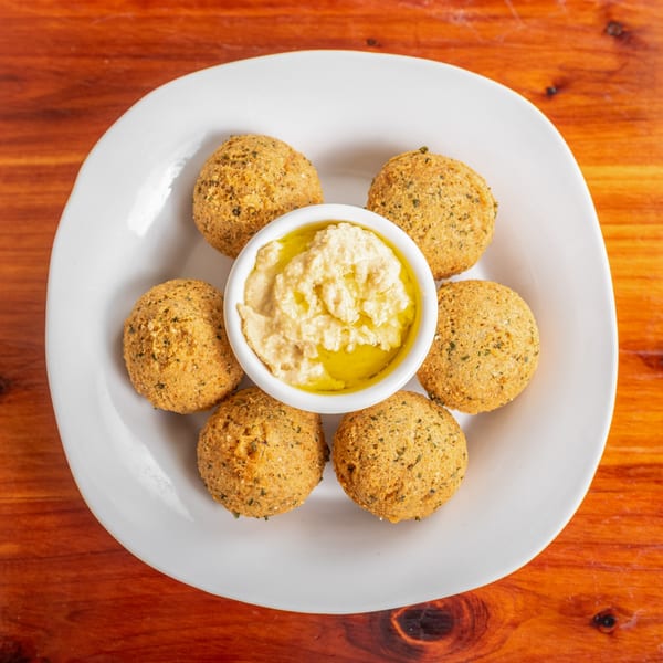 Six golden-brown falafel balls surround a white bowl of hummus drizzled with olive oil on a white plate.